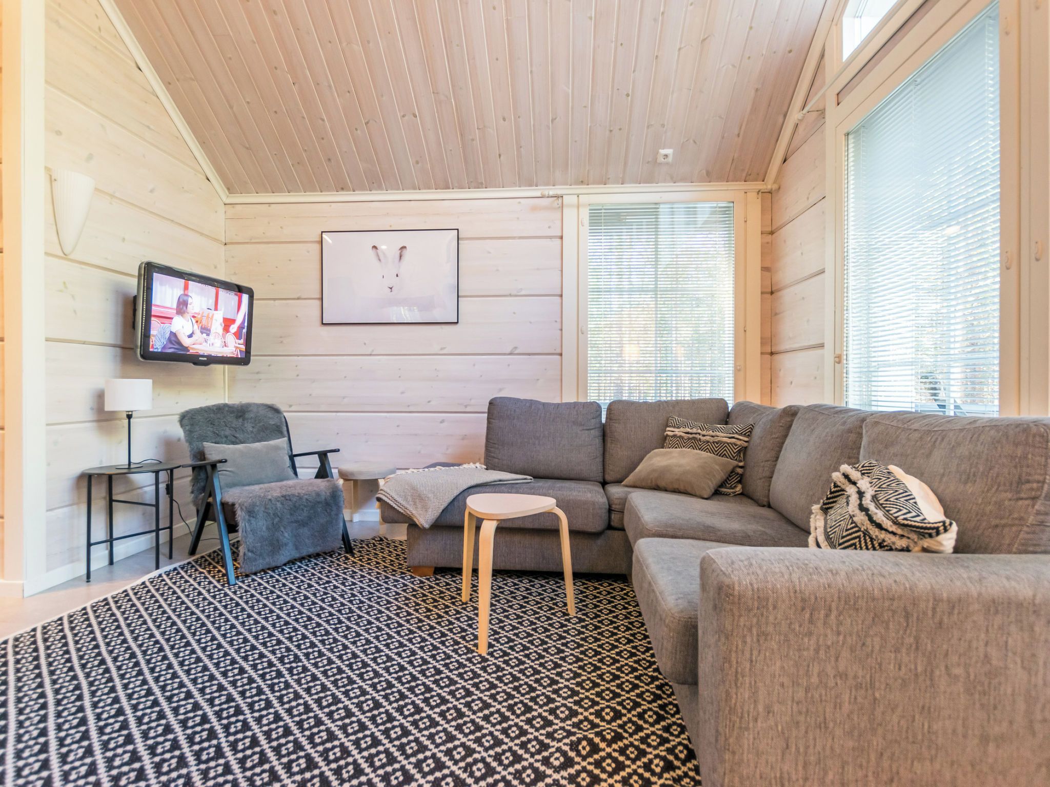 Un salon confortable avec un canapé, un fauteuil et une petite table. Un téléviseur est fixé au mur, et un tableau encadré est accroché au-dessus du canapé.
