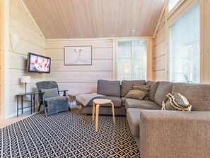Un salon confortable avec un canapé, un fauteuil et une petite table. Un téléviseur est fixé au mur, et un tableau encadré est accroché au-dessus du canapé.