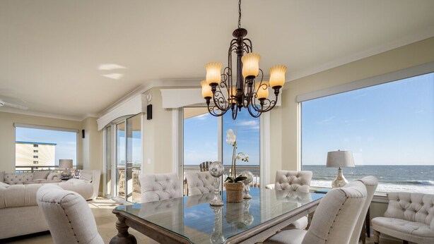 A spacious living room with a glass-top dining table surrounded by plush chairs, a decorative chandelier, and a view of the ocean. - Luxurious 4 bedroom oceanfront condo in the most prestigious building in OC, (Ocean City)