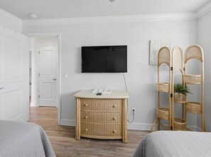 Una habitación con una televisión instalada en la pared, una mesita de mimbre, una silla de ratán y una cama.