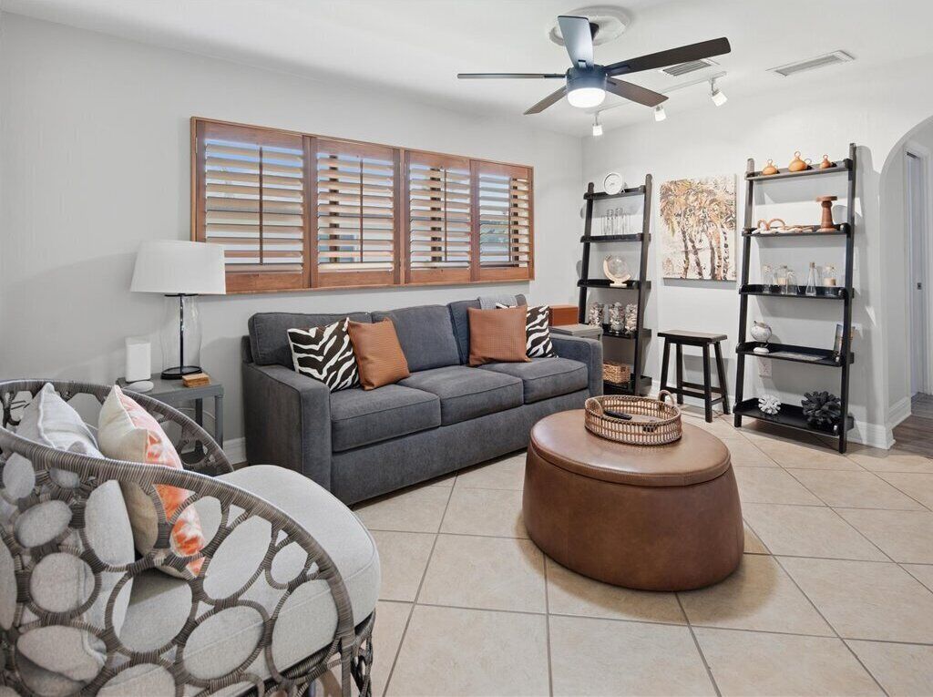 A living room with a grey sofa, patterned cushions, a round ottoman, a wicker chair, a ceiling fan, and a bookshelf with decorative items.
