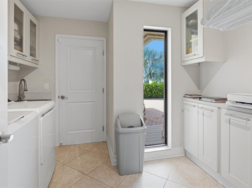 Une cuisine avec des meubles blancs, une porte, une poubelle et une fenêtre donnant sur un espace verdoyant.