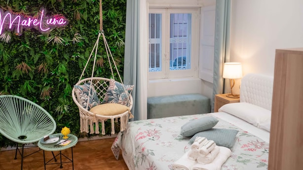 Una habitación acogedora con una silla colgante, una mesita con un pastel, una cama con ropa de cama floral y un letrero de neón que dice "Mare Luna".
