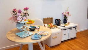Un pequeño comedor con una mesa lista para el desayuno, que incluye bollería y una taza de café. Hay dos arreglos florales sobre la mesa y un armario blanco con cajones al fondo.
