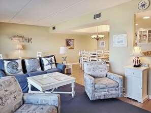 Una sala de estar con un sofá azul, sillones estampados, una mesa de centro blanca y un comedor con sillas y una mesa blancos.
