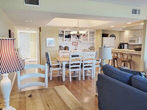 Uma sala de jantar com mesa de madeira e cadeiras brancas, uma cozinha com armários e eletrodomésticos brancos, e um sofá azul.