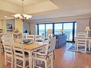 Uma sala de jantar com cadeiras brancas e uma mesa de madeira, uma sala de estar com sofás azuis e vista para o oceano através de grandes janelas.