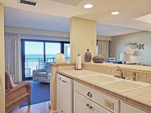 A hotel room with a balcony, a dining area, a kitchen with a sink, and a large window. - Oceanfront Condo in Gated Peninsula Locale, Elevator, Pools, Dock, 7-Mile Beach (Pawleys Island)