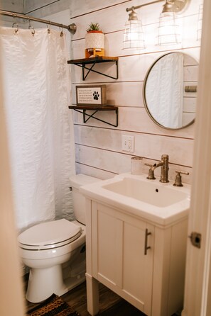 A bathroom with a toilet, sink, mirror, shower curtain, and wooden wall paneling. - Adorable Cottage on a Storybook Farm (Danielsville)