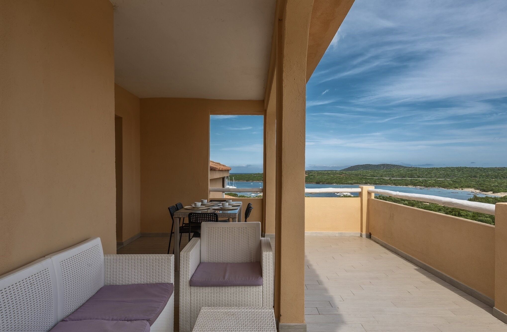 A balcony with a table set for dining, wicker furniture, and a view of a coastal landscape.