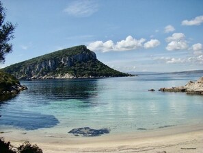Un paisaje costero con playa arenosa, mar tranquilo y una colina verde destacada con acantilados.
