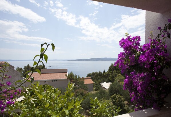 A balcony with a view of the sea and mountains, surrounded by greenery and purple flowers.