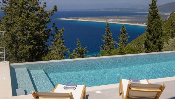 A pool with two lounge chairs, a clear view of the coastline, and surrounding greenery.