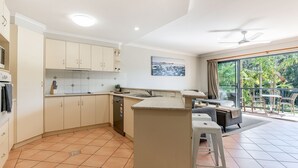 A modern kitchen with white cabinets, a built-in oven, and a dining area with a balcony overlooking greenery. - The Sails - 2-21 Beech Street (Evans Head)