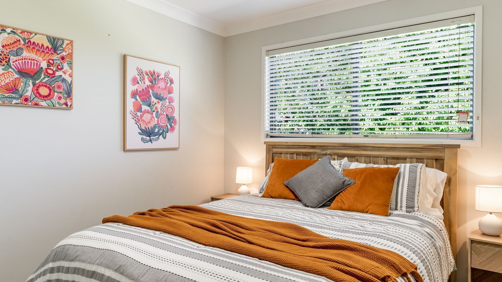 Un dormitorio con cama, mesita de noche, ventana con persianas y dos obras de arte florales en la pared.