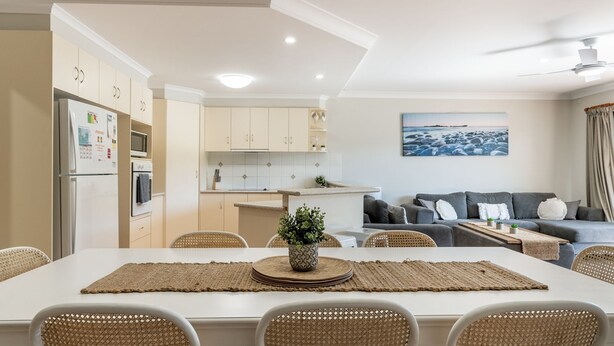 A modern kitchen with white cabinets, a dining area with a large table, and a living room with a grey sofa and a painting of a beach. - The Sails - 2-21 Beech Street (Evans Head)