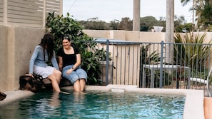 Two individuals sitting by a swimming pool with a concrete wall and metal railing in the background.