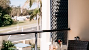 A balcony with a railing, a table with two glasses of a beverage, and a view of a street and greenery.
