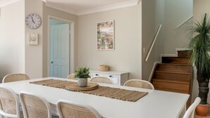 A dining area with a white table, woven placemats, wicker chairs, a clock, a framed picture, a small plant, and a staircase. - The Sails - 2-21 Beech Street (Evans Head)