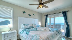 A bedroom with a bed, a nightstand, a ceiling fan, a window with blinds, and a view of the ocean through a balcony. - Oceanfront | Private Pool & Hot Tub | Adventures in Paradise (Oak Island)