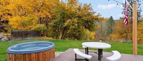 A hot tub, picnic table, and outdoor seating area on a wooden deck with a view of autumn trees.