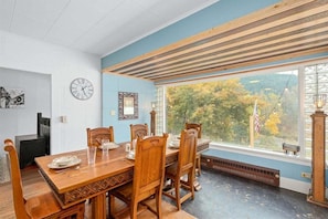 A dining area with a wooden table and chairs, a clock on the wall, and a view of trees through a large window. - Downtown Leavenworth Cottage w/ Hot Tub, Fireplace, BBQ (Leavenworth)