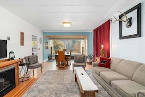 A living room with a fireplace, a sofa, a dining table with chairs, and a large window. - Downtown Leavenworth Cottage w/ Hot Tub, Fireplace, BBQ (Leavenworth)