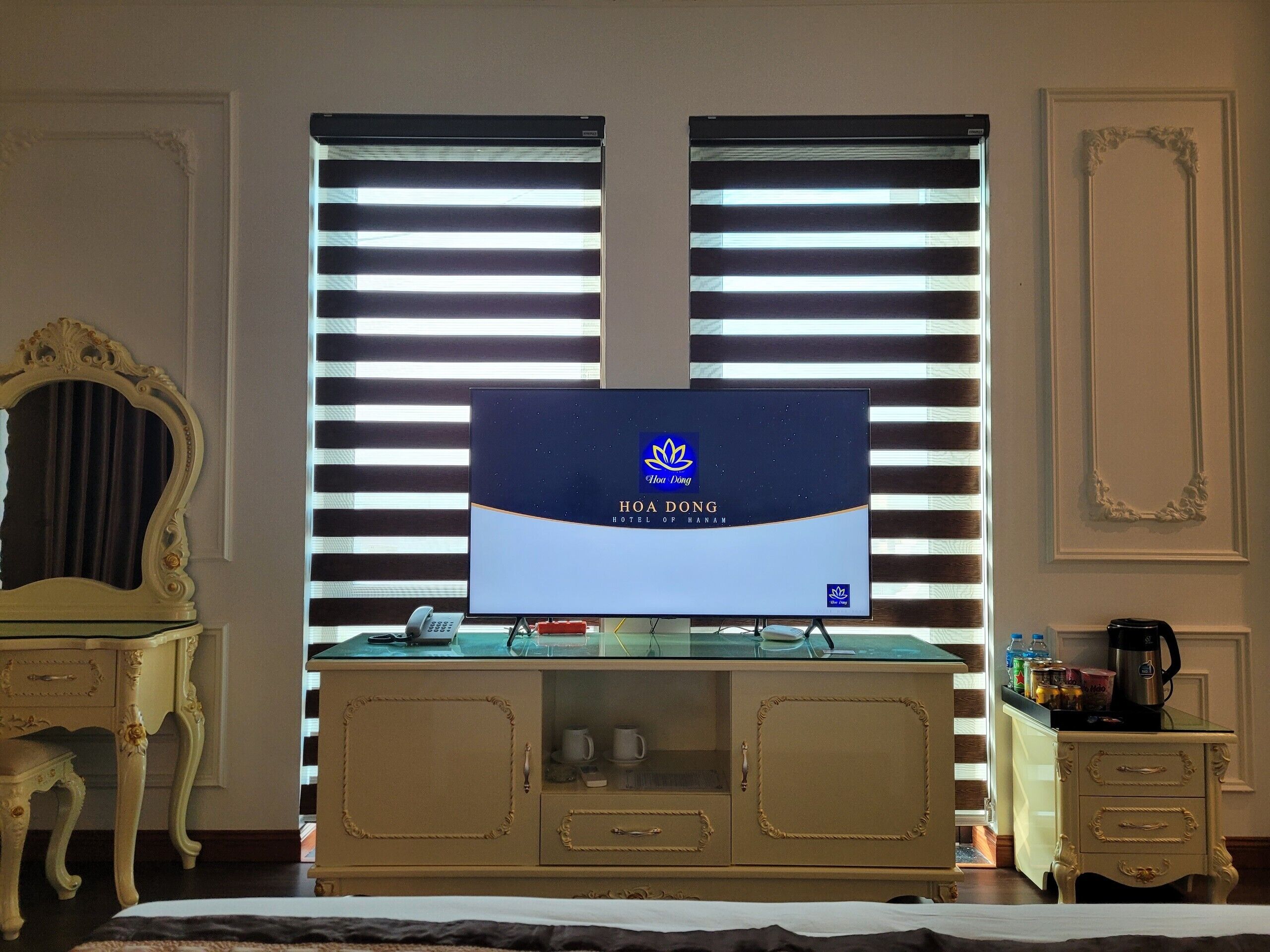 A hotel room with a large TV screen displaying the hotel's name, a minibar, a desk with a telephone, and a decorative mirror.