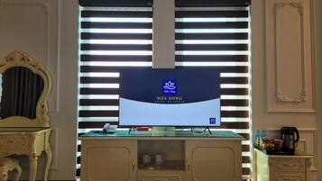 A hotel room with a large TV screen displaying the hotel's name, a minibar, a desk with a telephone, and a decorative mirror.