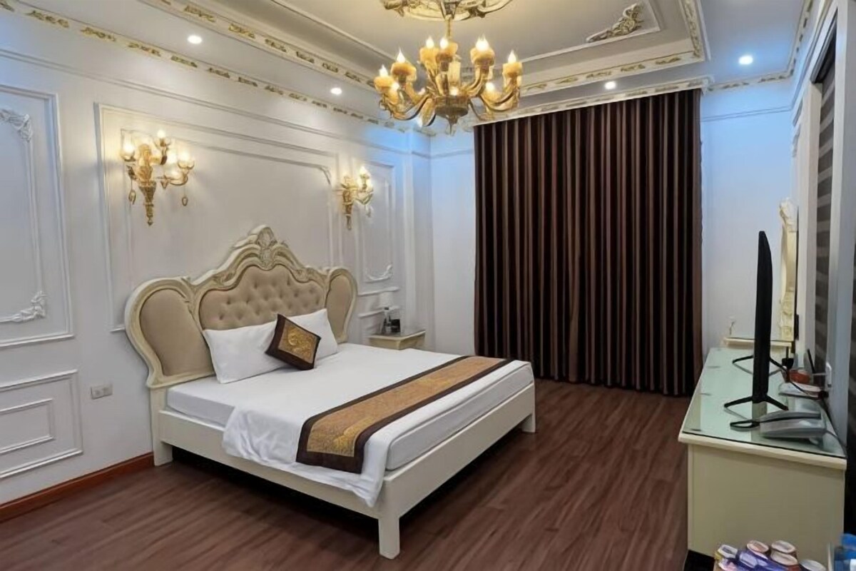 A bedroom with a large bed, wall sconces, a chandelier, a desk with a lamp, and a window with curtains.