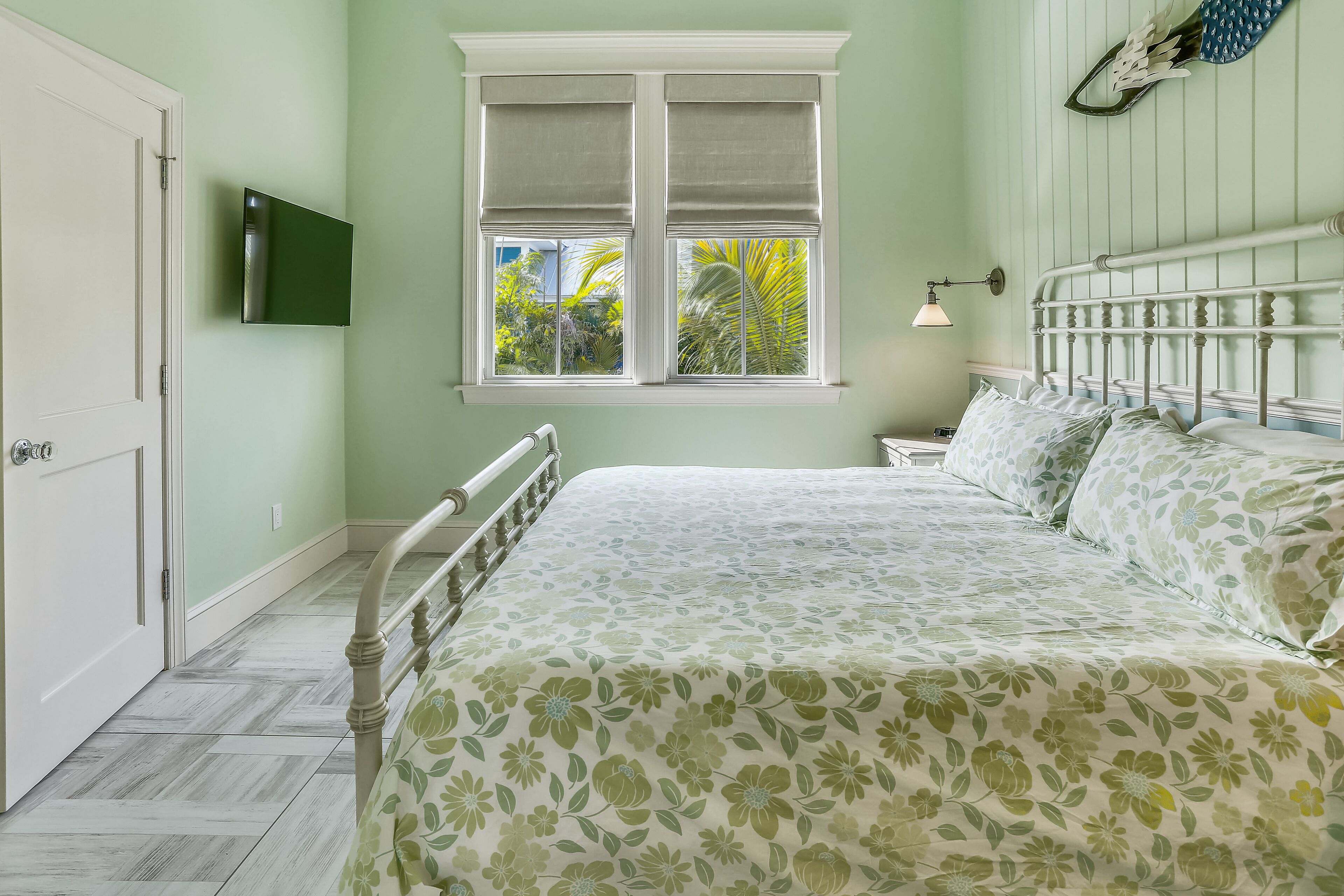 A bedroom with a bed, a window with blinds, a wall-mounted TV, and a nightstand with a lamp.