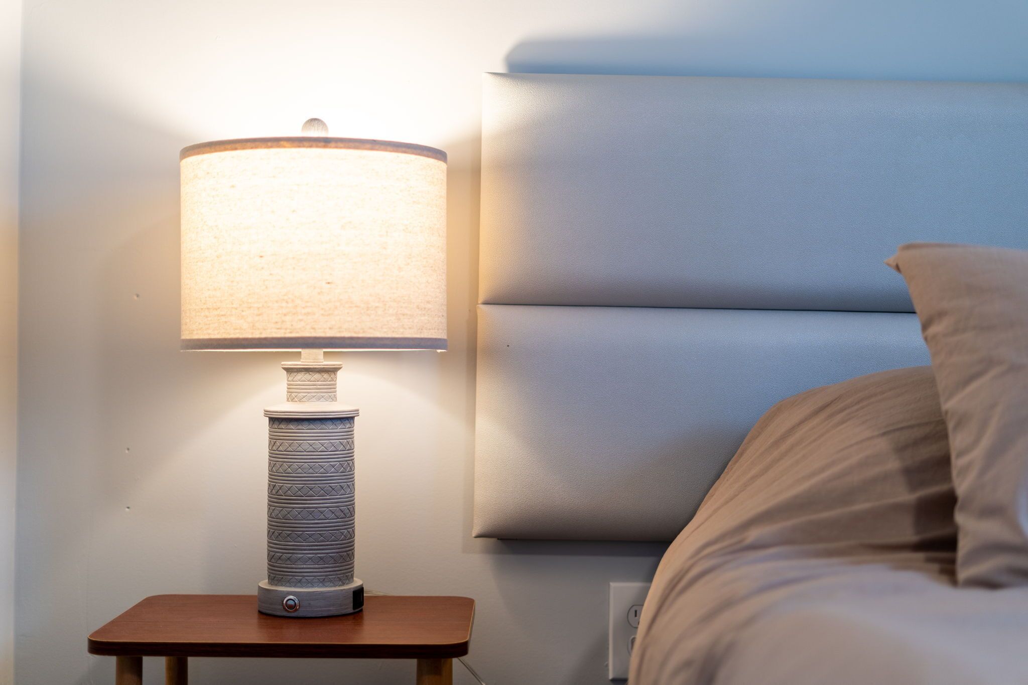 Ein Nachttisch mit Lampe, ein Bett mit Kopfteil und eine Steckdose an der Wand.