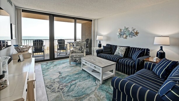 Een ruime woonkamer met een groot balkon met uitzicht op het strand, ingericht met gestreepte banken, fauteuils en een salontafel.