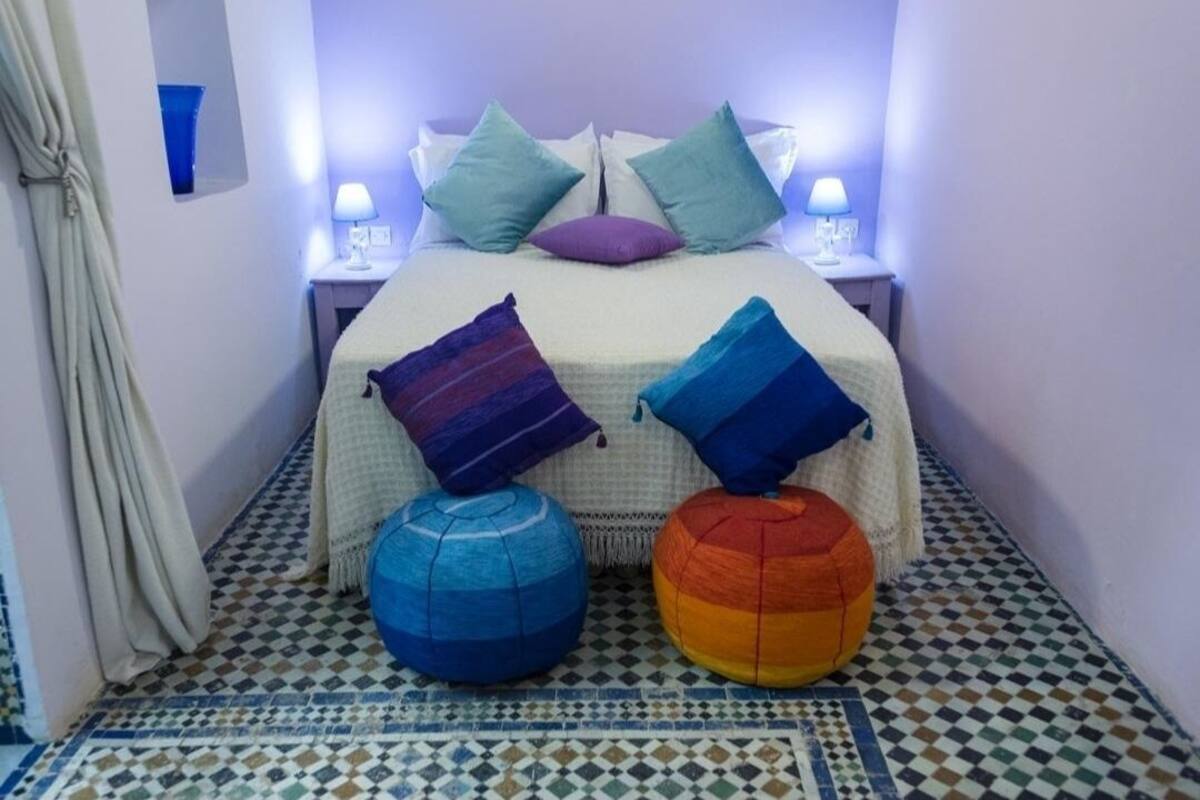 A bedroom with a bed, two nightstands, a lamp, a purple pillow, and two colorful ottomans.
