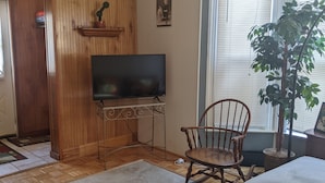 A living room with a wooden floor, a round coffee table, a television on a stand, a rocking chair, a sofa, a potted plant, and a window with blinds.