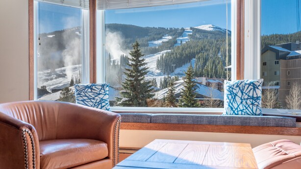 A room with a large window offering a view of a snowy mountain landscape, a leather sofa, and a wooden table. - Amazing Copper Mountain Views, free wifi, free parking. (Copper Mountain)