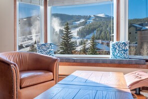 A room with a large window offering a view of a snowy mountain landscape, a leather sofa, and a wooden table. - Amazing Copper Mountain Views, free wifi, free parking. (Copper Mountain)