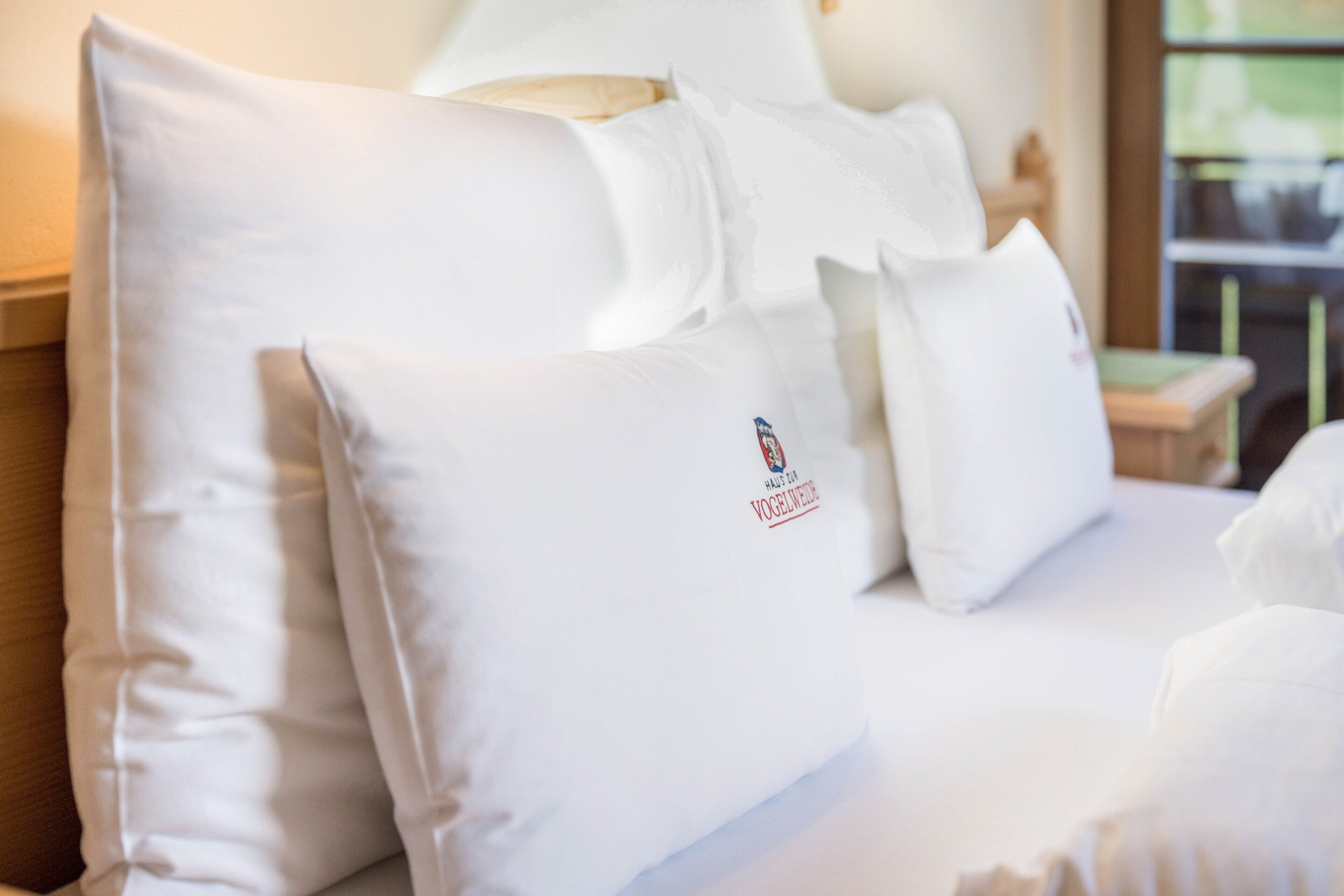 Ett hotellrum med prydligt placerade vita kuddar, en tydlig logotyp på en av kuddarna och ett fönster i bakgrunden.
