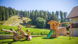 Leikkikenttä, jossa on liukumäki, puinen rakennelma ja leikkimökki maaseutumaisessa ympäristössä.