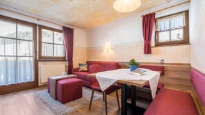 Une pièce chaleureuse avec un plancher en bois, un canapé rouge, une petite table recouverte d’une nappe blanche et une fenêtre avec des rideaux.