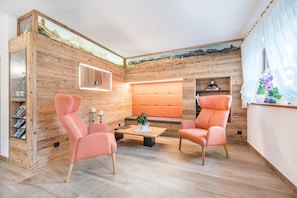 A room with wooden walls, a cushioned bench, two pink chairs, a small table with a plant, and a shelf with books and a decorative item.
