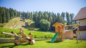 A playground with a slide and climbing structures, surrounded by a grassy area and a forested hill.