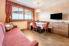 A cozy room with a sofa, a dining table with chairs, a TV mounted on the wall, and a window with curtains.