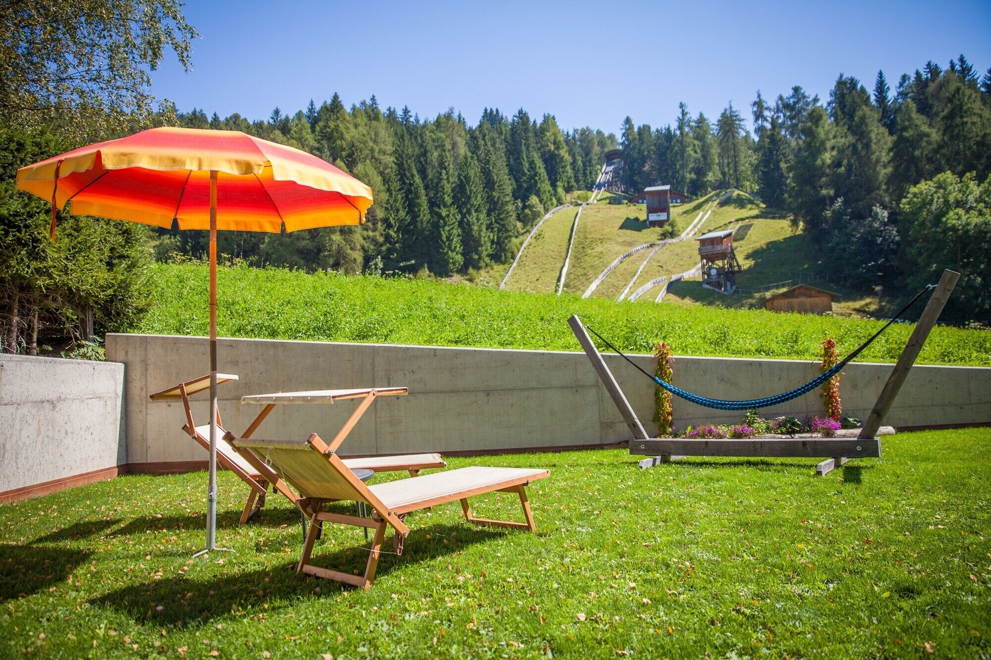Un espace extérieur verdoyant avec un hamac, un fauteuil de relaxation et un parasol rouge et jaune.