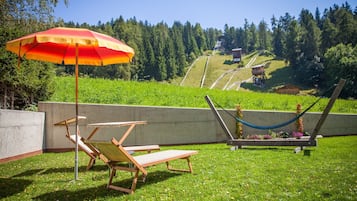 Un espace extérieur verdoyant avec un hamac, un fauteuil de relaxation et un parasol rouge et jaune.