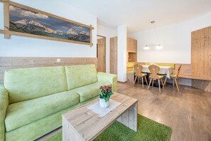 A modern living room with a green sofa, a wooden coffee table, and a dining area with wooden chairs and a table.