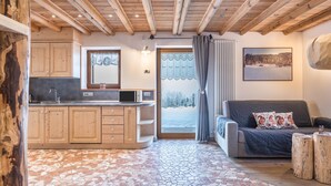 Une pièce chaleureuse avec un plafond en bois, un coin cuisine avec des placards, un canapé avec des coussins et une vue sur l’extérieur, visible à travers une porte.
