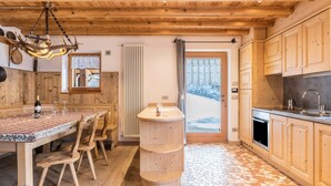 Un intérieur en bois comprenant une table à manger, des chaises, un lustre, un coin cuisine et une vue sur l’extérieur, visible à travers une porte.