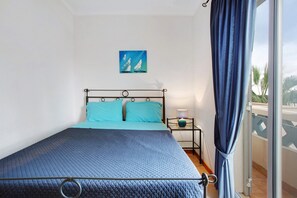 Een bed met een blauwe sprei, een klein nachtkastje met een lamp, een aan de muur hangend schilderij en uitzicht door een raam met gordijnen.