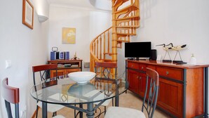 Un comedor con una mesa de vidrio, sillas, una escalera espiral de madera, un televisor y un mueble con adornos.